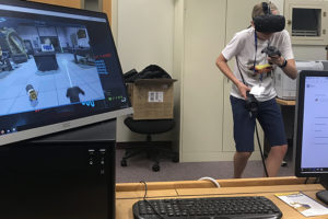 A student wears a virtual reality headset, and a computer monitor displays what the student sees.
