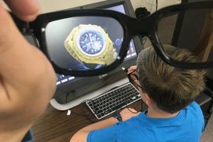 A student works at a computer, and, through glasses, an image of a large gold watch is seen.